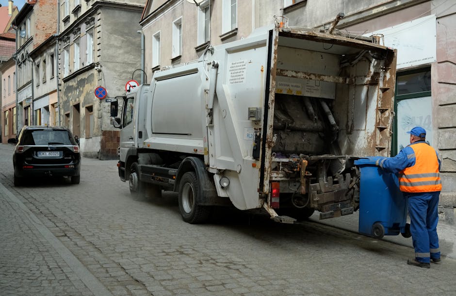 Two waste collection workers are engaged in rubbish removal activities on a city street during early evening or dusk. One worker, wearing a dark uniform with reflective orange and yellow stripes on th